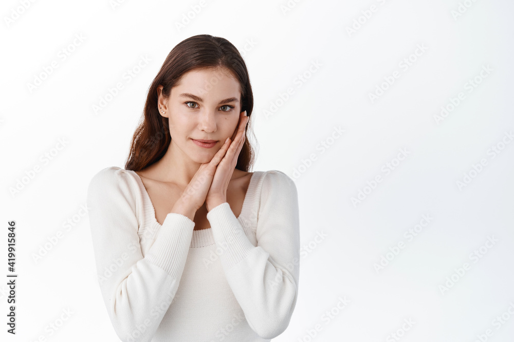 Attractive girlfriend can not stop gazing at you. Slender caucasian woman tenderly touching cheek with palm and smiling cheerfully at camera while standing over white wall Attractive girlfriend can not stop gazing at you. Slender caucasian woman tenderly touching cheek with palm and smiling cheerfully at camera while standing over white wall