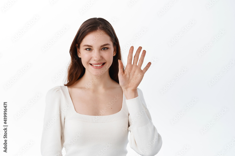 Young woman smiling and saluting, waving hand to say hello, greeting friends, say hi and welcome, goodbye gesture, standing against white background Young woman smiling and saluting, waving hand to say hello, greeting friends, say hi and welcome, goodbye gesture, standing against white background