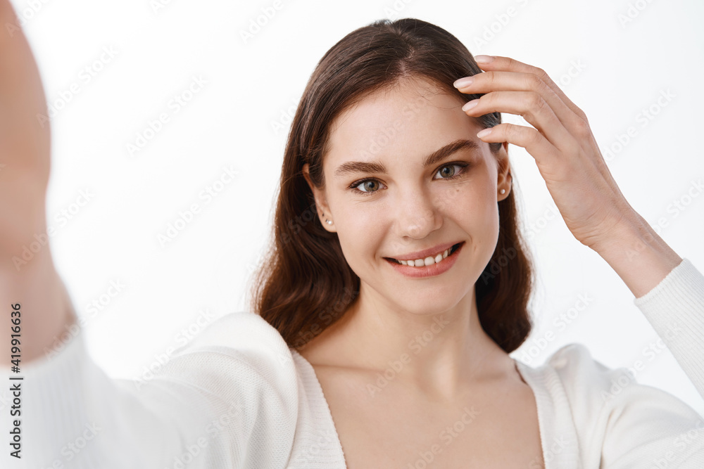 Attractive young woman holding camera in stretched hand and taking selfie, making her photo on mobile phone, smiling, standing over white background Attractive young woman holding camera in stretched hand and taking selfie, making her photo on mobile phone, smiling, standing over white background