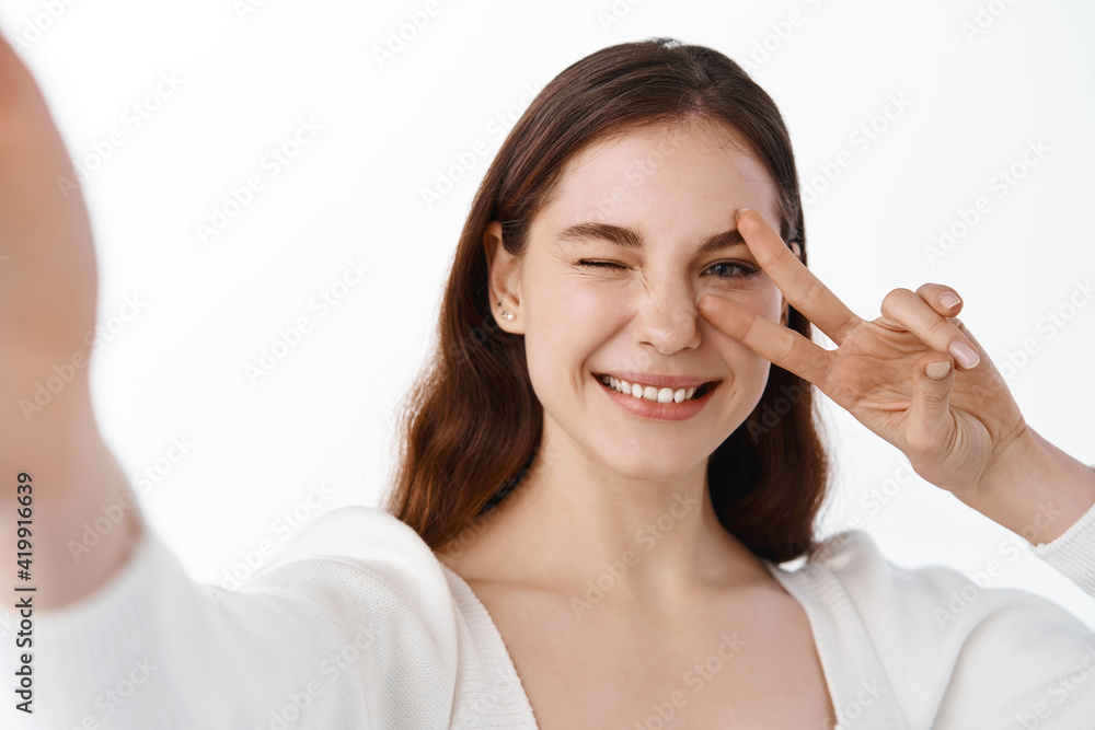Attractive happy woman taking selfie with v-sign, stretch out hand to hold camera, making positive face, smile and peace gesture, standing over white background Attractive happy woman taking selfie with v-sign, stretch out hand to hold camera, making positive face, smile and peace gesture, standing over white background