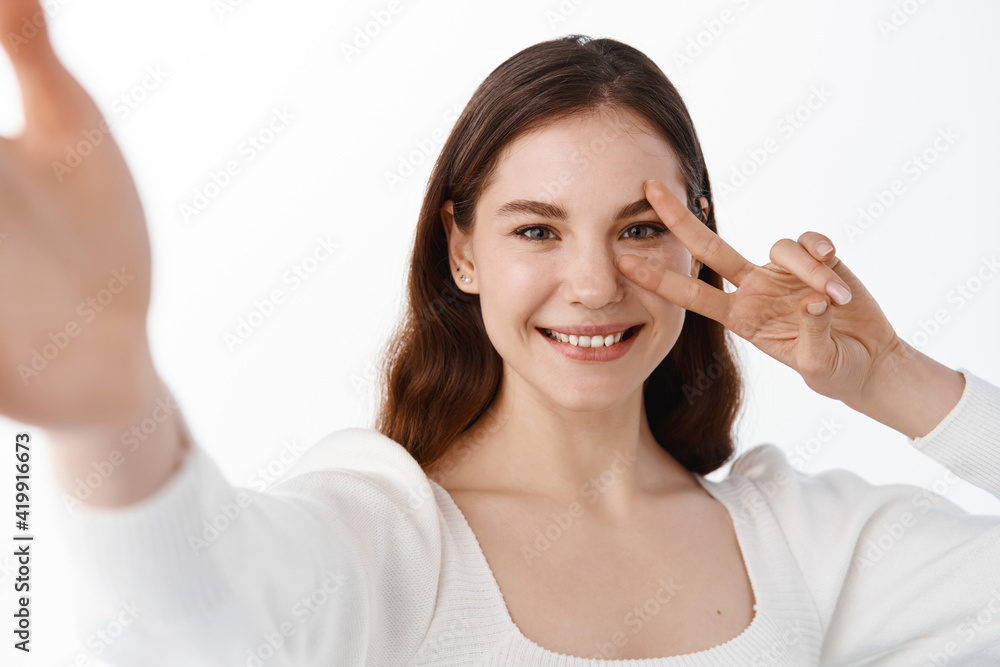 Young beautiful girl student taking selfie on mobile camera, holding phone with stretch out hand, showing v-sign and smiling, standing over white background Young beautiful girl student taking selfie on mobile camera, holding phone with stretch out hand, showing v-sign and smiling, standing over white background