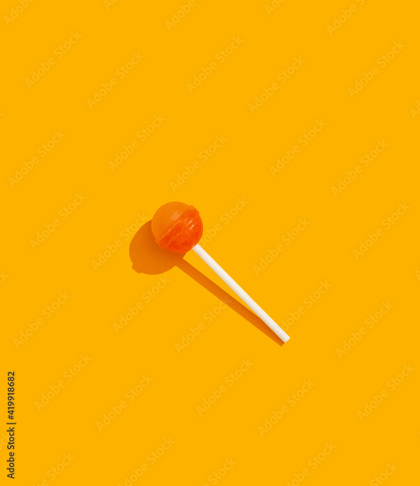 Orange and yellow hard candy lollipop on stick laying on orange background top view with trendy hard shadows. Summer, leisure, vacation minimal concept Orange and yellow hard candy lollipop on stick laying on orange background top view with trendy hard shadows. Summer, leisure, vacation minimal concept