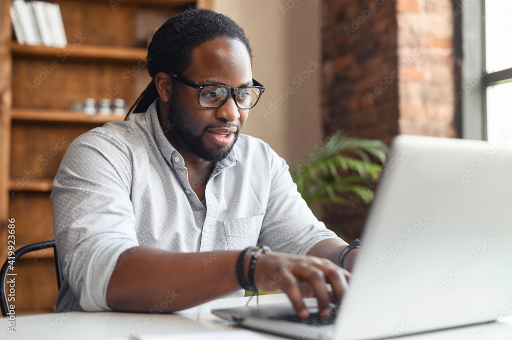 Focused African-American man wearing stylish eyeglasses and smart casual shirt using a laptop for work sitting in the modern office space, a multiracial office employee answering emails, typing Focused African-American man wearing stylish eyeglasses and smart casual shirt using a laptop for work sitting in the modern office space, a multiracial office employee answering emails, typing