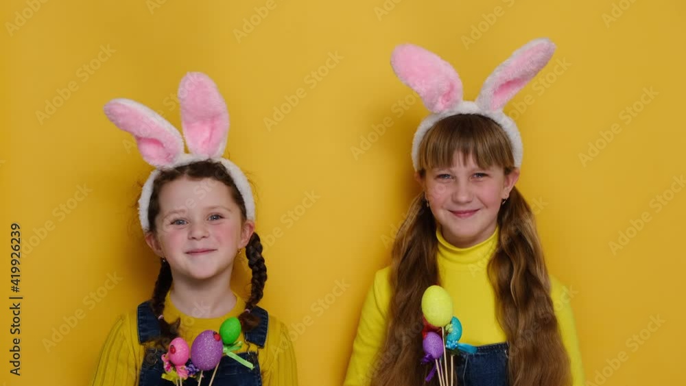 Portrait of happy cute two adorable little sisters holding small colored eggs, being symbol of religious holiday, wearing bunny ears, isolated on yellow studio background. Easter celebration concept Portrait of happy cute two adorable little sisters holding small colored eggs, being symbol of religious holiday, wearing bunny ears, isolated on yellow studio background. Easter celebration concept