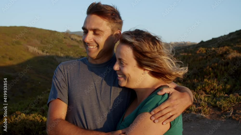 Happy couple of peaceful tourists in love enjoying leisure time outdoors, standing at cliff at sunset, hugging and talking, enjoying breathtaking view of sea. Travel activity or romance concept Happy couple of peaceful tourists in love enjoying leisure time outdoors, standing at cliff at sunset, hugging and talking, enjoying breathtaking view of sea. Travel activity or romance concept