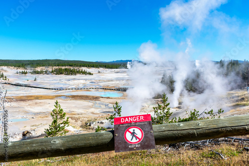 Danger Hazardous Thermal Ground warning sign. Blurred scenic landscape with boardwalk among pools and active geysers of Norris Geyser Basin in Yellowstone National Park.