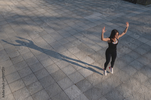Sporty woman runner in training on grey sidewalk . Photo of attractive woman in fashionable sportswear. Dynamic movement. Top view. Sun spot. Sport and healthy lifestyle. Place for text.