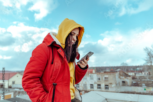 Portrait of young woman with mobile phone on roof of building. Brunette in yellow hoodie and red jacket browsing smartphone in cloudy weather.