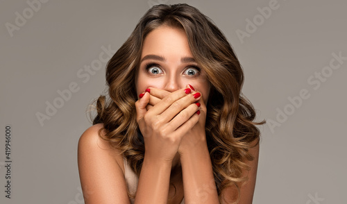 Shocked and surprised girl screaming covering mouth her hands.Curly hair woman amazed.Beautiful girl with curly hairstyle and red nails manicure.Presenting your product.Expressive facial expressions