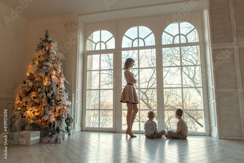 Mom with two little boys standing at the window in a big house next to the Christmas tree