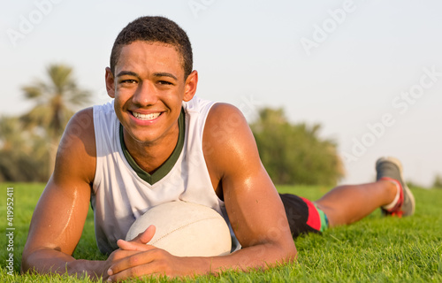 Happy fit black teen athlete resting on the grass with a rugby ball. Sports lifestyle.