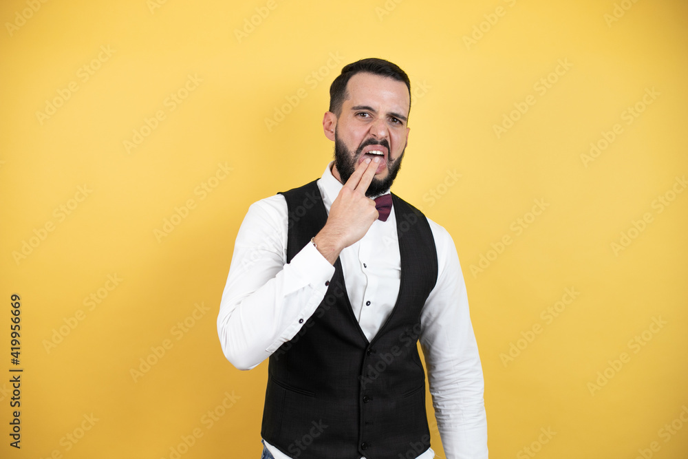 Young man with beard wearing bow tie and vest disgusted with his hand inside his mouth Young man with beard wearing bow tie and vest disgusted with his hand inside his mouth