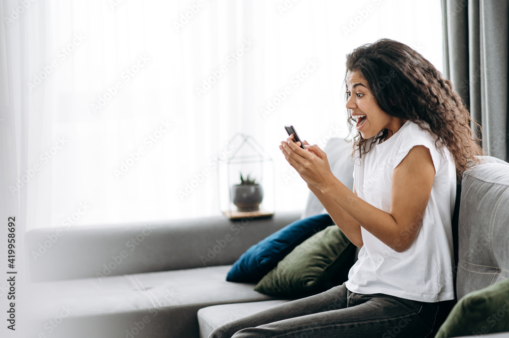 Happy female freelancer is overjoyed. Joyful young African American woman sits on the sofa, use smartphone, got a good news, joyfully looks at the phone screen Happy female freelancer is overjoyed. Joyful young African American woman sits on the sofa, use smartphone, got a good news, joyfully looks at the phone screen