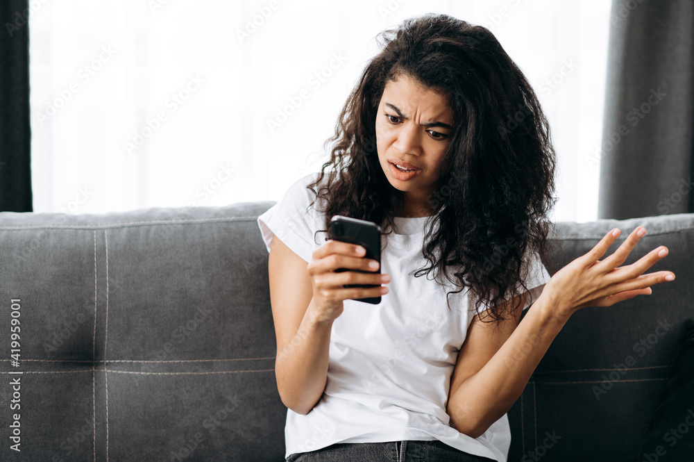 Upset shocked young woman, confused looks at the phone screen. Beautiful african american female got an unexpected message or news, sits on the couch and browsing the internet or messages Upset shocked young woman, confused looks at the phone screen. Beautiful african american female got an unexpected message or news, sits on the couch and browsing the internet or messages
