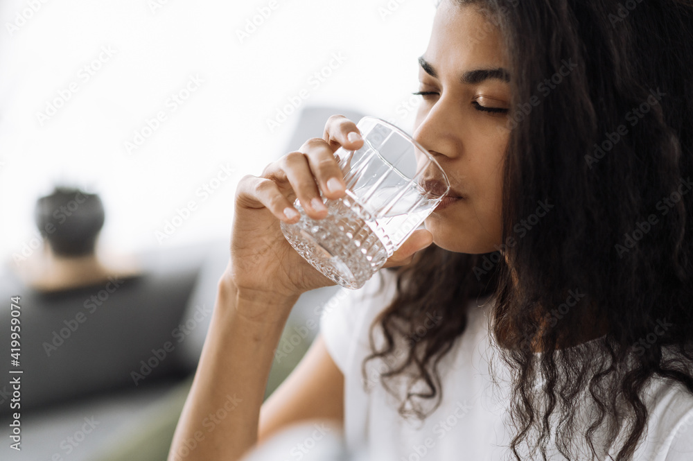 Healthy lifestyle. African american young woman drinks a glass of water while sitting on the sofa at home. Beautiful healthy woman follow healthy lifestyle Healthy lifestyle. African american young woman drinks a glass of water while sitting on the sofa at home. Beautiful healthy woman follow healthy lifestyle