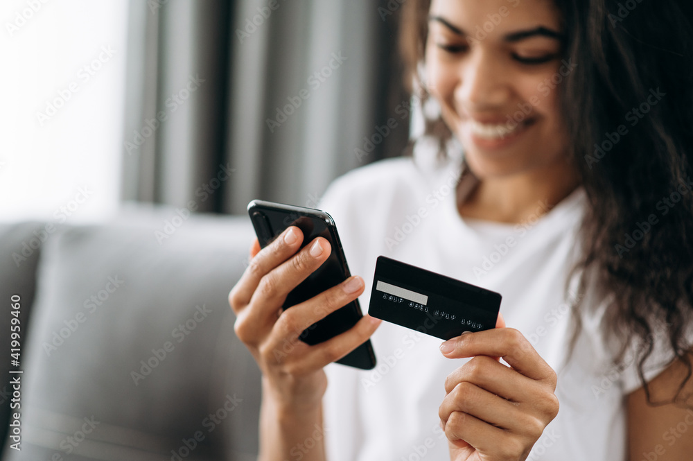 A credit card and smartphone in female hands. Joyful african american young woman in defocus looks at the phone screen, shopping online. Credit card and phone in the focus A credit card and smartphone in female hands. Joyful african american young woman in defocus looks at the phone screen, shopping online. Credit card and phone in the focus