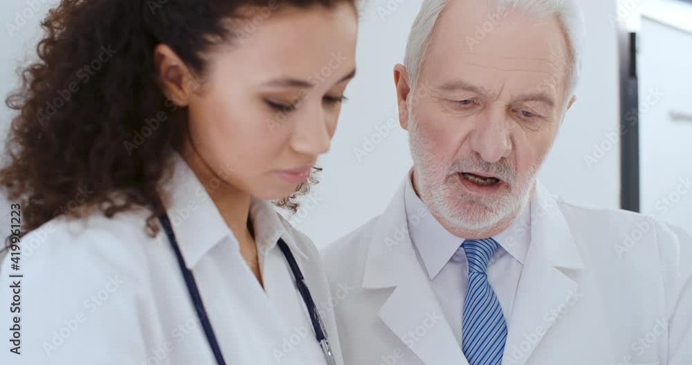 Close-up portrait two medical workers aged caucasian man and young curly hair african american woman, doctors discussing medical case, talking about health report while standing in hospital Close-up portrait two medical workers aged caucasian man and young curly hair african american woman, doctors discussing medical case, talking about health report while standing in hospital