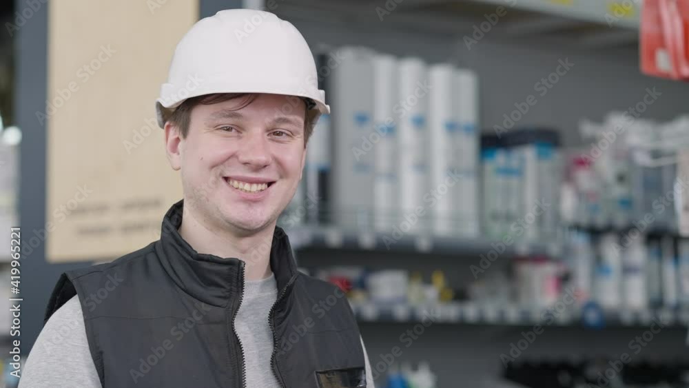 Close-up portrait of young positive Caucasian man in hard hat posing in hardware store with blurred shelves at background. Happy worker looking at camera smiling. Service and occupation. Close-up portrait of young positive Caucasian man in hard hat posing in hardware store with blurred shelves at background. Happy worker looking at camera smiling. Service and occupation.