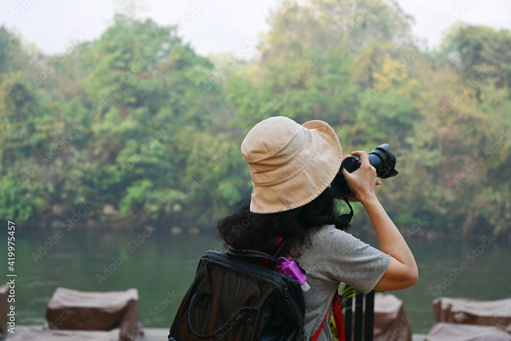 Obraz premium Asian female tourists wearing a cream-colored hat, carrying black backpack, hanging bottles of purple hand sanitizer, taking photos of the nature of the beautiful forest and river on a camera vacation