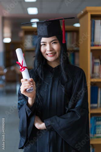 A young happy Asian woman university graduates in graduation gown and cap wears a face mask holds a degree certificate to celebrate her education achievement on the commencement day. Stock photo