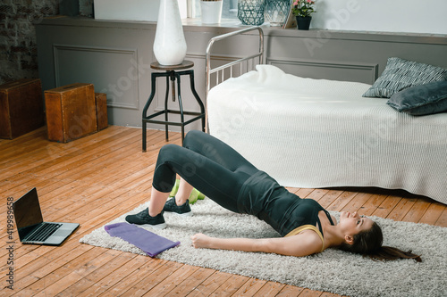 A young woman goes in for sports at home, online workout . The athlete  pumps up the buttocks, makes the gluteal bridge in the bedroom, in the background there is a bed, a vase, a carpet. photo