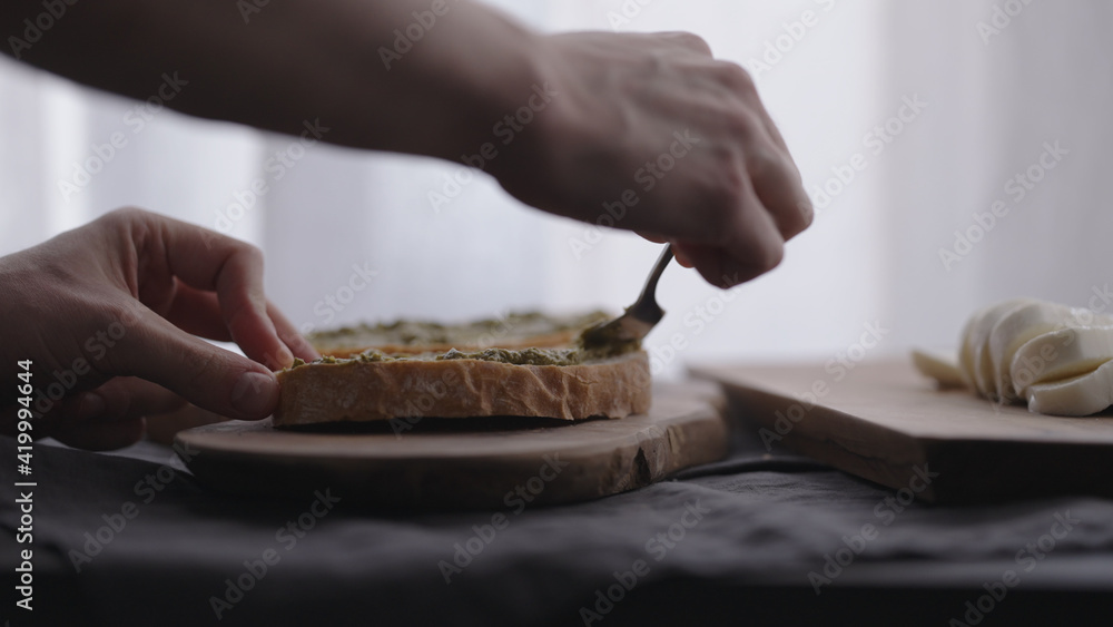 man making ciabatta bruschettas with mozzarella and pesto on olive board with window on backgorund