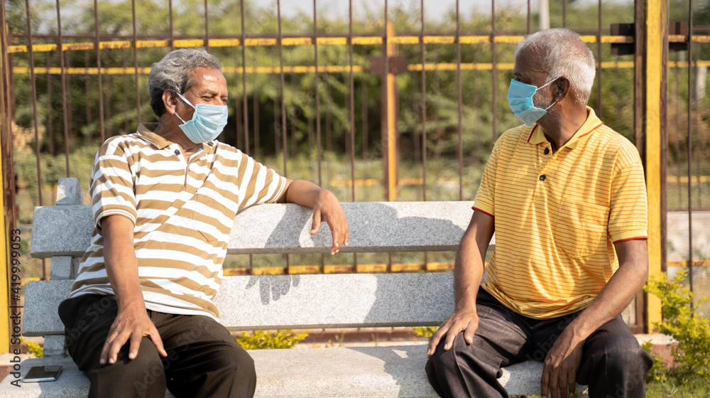 Two old senior friends with medical mask spending good time by talking and while maintaining social distancing at park - concept of new normal lifestyle due to coronavirus covid-19 pandemic