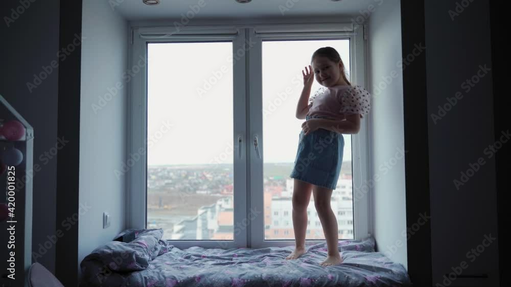 Сhildhood. Cheerful attractive girl is dancing at the window. She smiles and laughs. She expresses herself. The city is visible outside the window Сhildhood. Cheerful attractive girl is dancing at the window. She smiles and laughs. She expresses herself. The city is visible outside the window