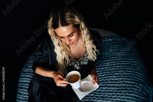 Blonde Frau mit Kaffee in der Hand, Neglische, Bett, dunkler Hintergrund, gestrickte graue Decke weiße Tasse mit Kaffee, Kaffee zum Frühstück, Abend, im Bett