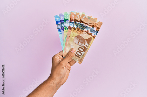 Hispanic hand holding canadian dollars banknotes over isolated pink background.