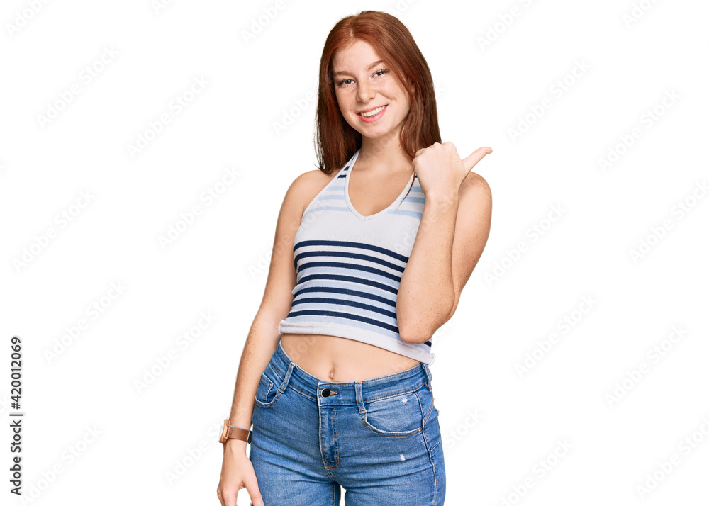 Young read head woman wearing casual clothes smiling with happy face looking and pointing to the side with thumb up. Young read head woman wearing casual clothes smiling with happy face looking and pointing to the side with thumb up.