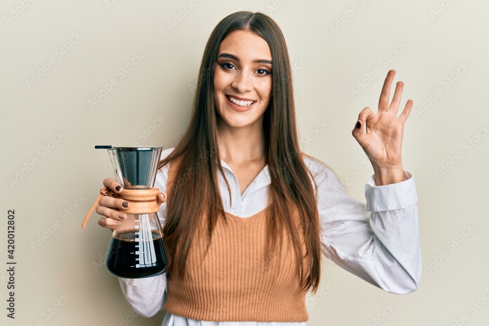 Beautiful brunette young woman holding soluble coffee maker doing ok sign with fingers, smiling friendly gesturing excellent symbol Beautiful brunette young woman holding soluble coffee maker doing ok sign with fingers, smiling friendly gesturing excellent symbol