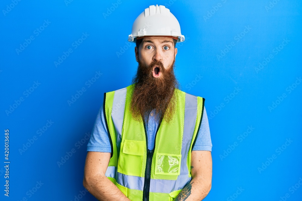 Redhead man with long beard wearing safety helmet and reflective jacket afraid and shocked with surprise expression, fear and excited face. Redhead man with long beard wearing safety helmet and reflective jacket afraid and shocked with surprise expression, fear and excited face.