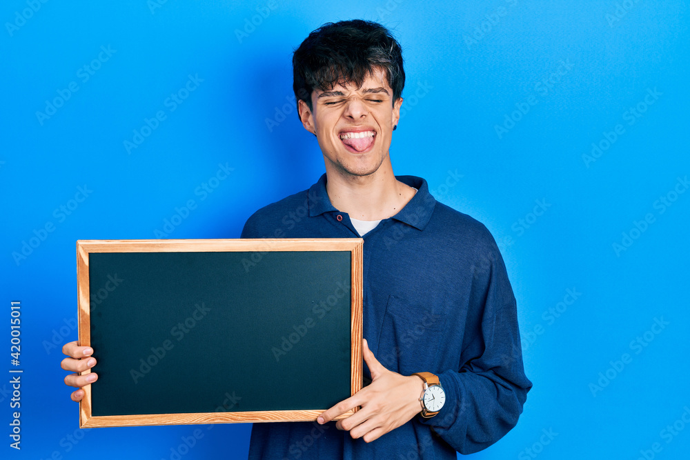 Handsome hipster young man holding blackboard sticking tongue out happy with funny expression. Handsome hipster young man holding blackboard sticking tongue out happy with funny expression.