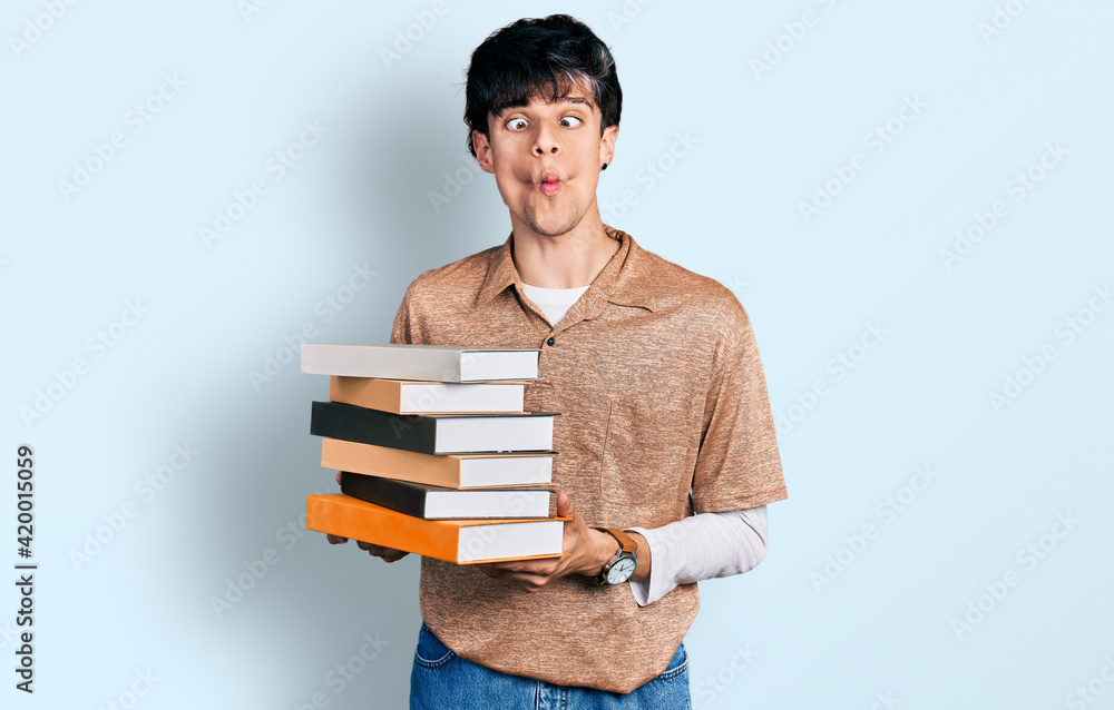 Handsome hipster young man holding a pile of books making fish face with mouth and squinting eyes, crazy and comical. Handsome hipster young man holding a pile of books making fish face with mouth and squinting eyes, crazy and comical.