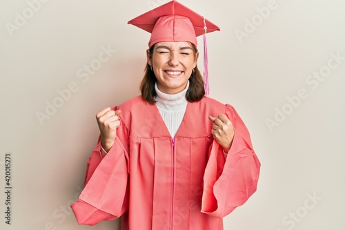 Young caucasian woman wearing graduation cap and ceremony robe very happy and excited doing winner gesture with arms raised, smiling and screaming for success. celebration concept.