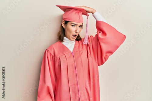 Young caucasian woman wearing graduation cap and ceremony robe surprised with hand on head for mistake, remember error. forgot, bad memory concept.