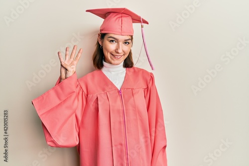 Young caucasian woman wearing graduation cap and ceremony robe showing and pointing up with fingers number four while smiling confident and happy.