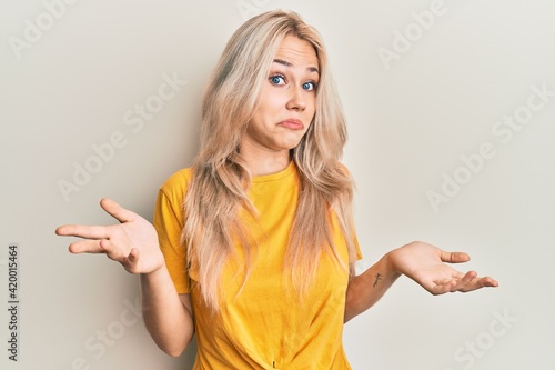 Beautiful caucasian blonde girl wearing casual tshirt clueless and confused expression with arms and hands raised. doubt concept.
