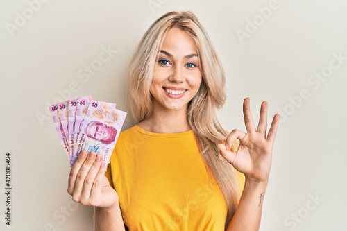 Beautiful caucasian blonde girl holding 50 mexican pesos banknotes doing ok sign with fingers, smiling friendly gesturing excellent symbol