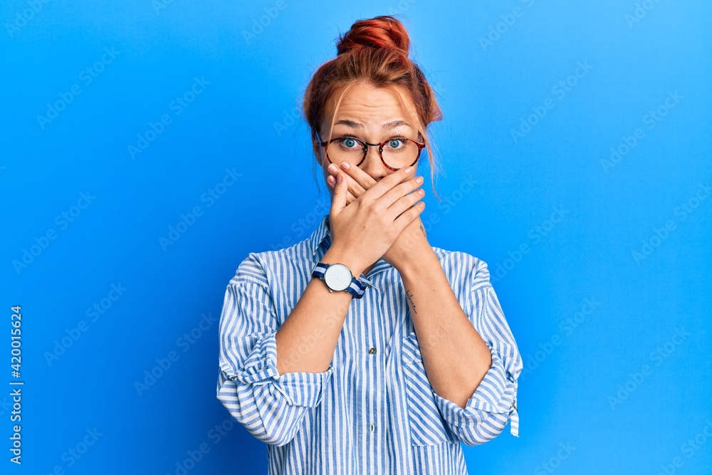 Young beautiful redhead woman wearing casual clothes and glasses over blue background shocked covering mouth with hands for mistake. secret concept. Young beautiful redhead woman wearing casual clothes and glasses over blue background shocked covering mouth with hands for mistake. secret concept.