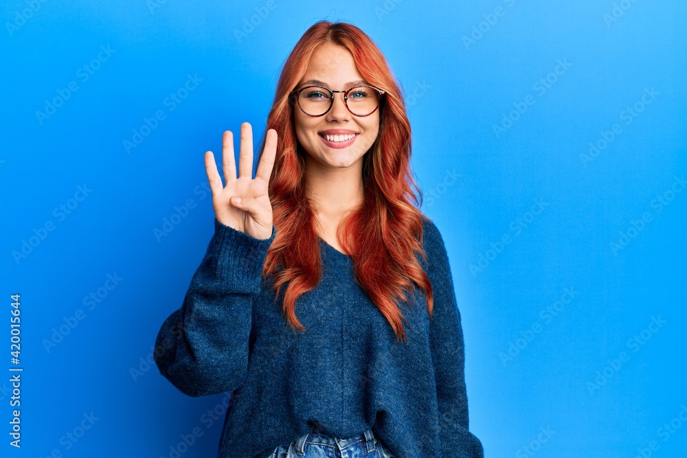 Young beautiful redhead woman wearing casual sweater and glasses over blue background showing and pointing up with fingers number four while smiling confident and happy.