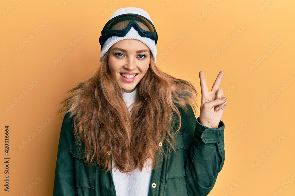 Beautiful blonde caucasian woman wearing snow wear and sky glasses smiling with happy face winking at the camera doing victory sign. number two. Beautiful blonde caucasian woman wearing snow wear and sky glasses smiling with happy face winking at the camera doing victory sign. number two.