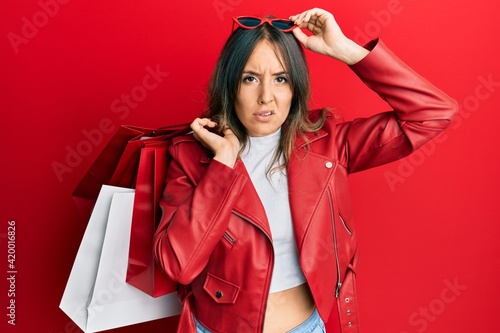 Young brunette woman holding shopping bags in shock face, looking skeptical and sarcastic, surprised with open mouth