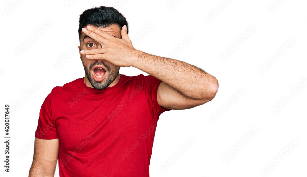 Hispanic man with beard wearing casual red t shirt peeking in shock covering face and eyes with hand, looking through fingers with embarrassed expression. Hispanic man with beard wearing casual red t shirt peeking in shock covering face and eyes with hand, looking through fingers with embarrassed expression.