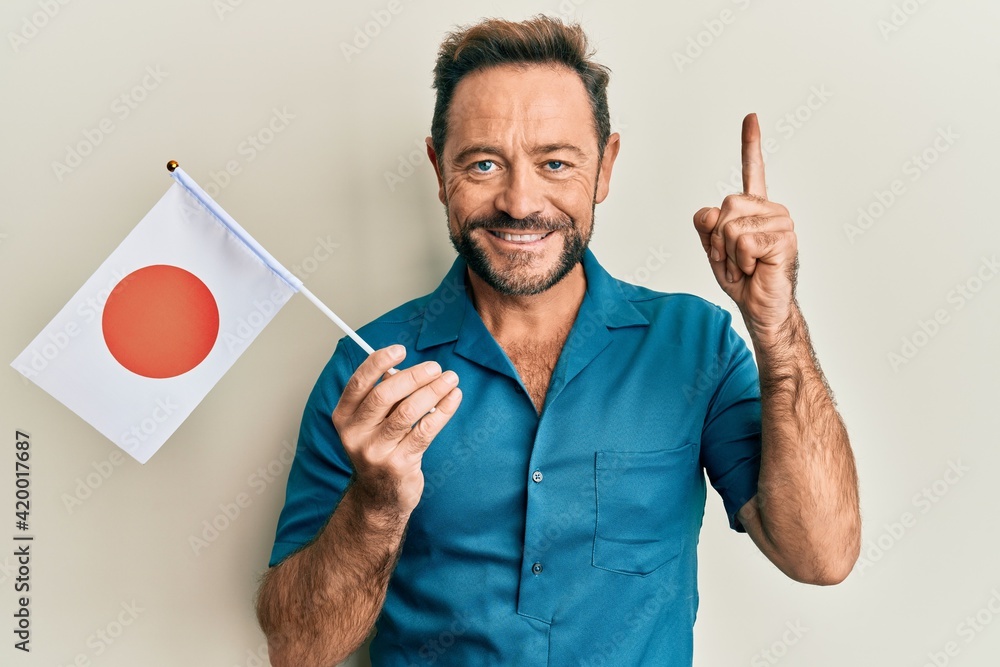 Middle age man holding japan flag smiling with an idea or question pointing finger with happy face, number one Middle age man holding japan flag smiling with an idea or question pointing finger with happy face, number one