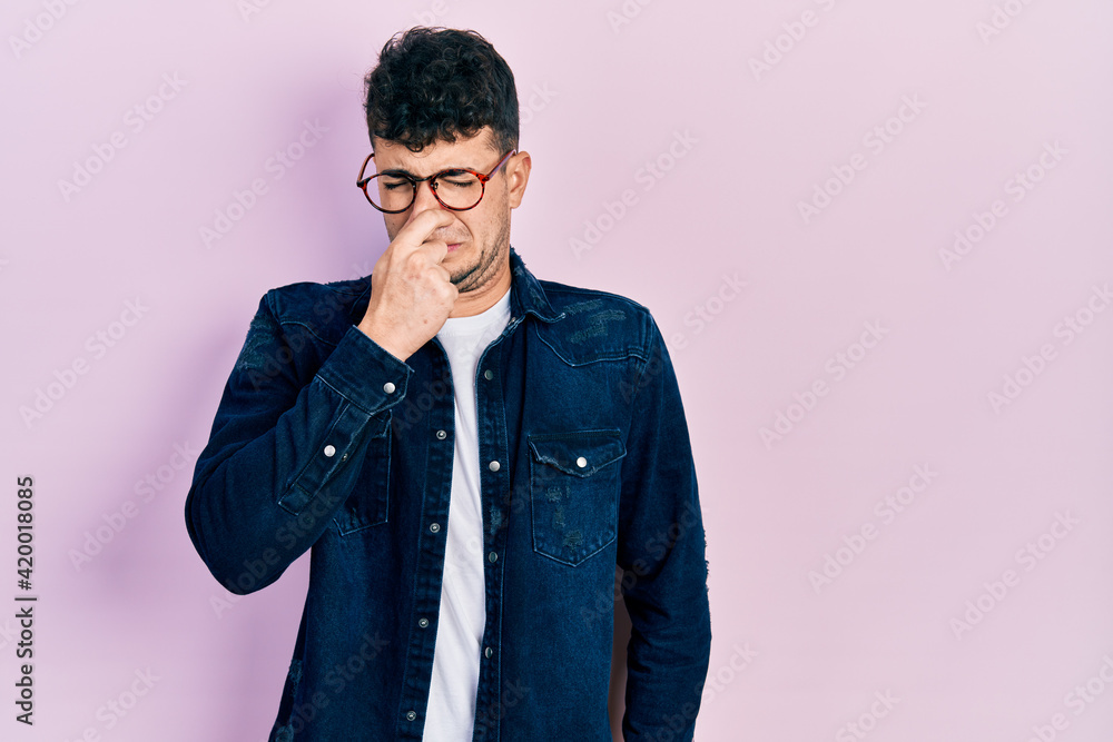 Young hispanic man wearing casual clothes and glasses smelling something stinky and disgusting, intolerable smell, holding breath with fingers on nose. bad smell Young hispanic man wearing casual clothes and glasses smelling something stinky and disgusting, intolerable smell, holding breath with fingers on nose. bad smell
