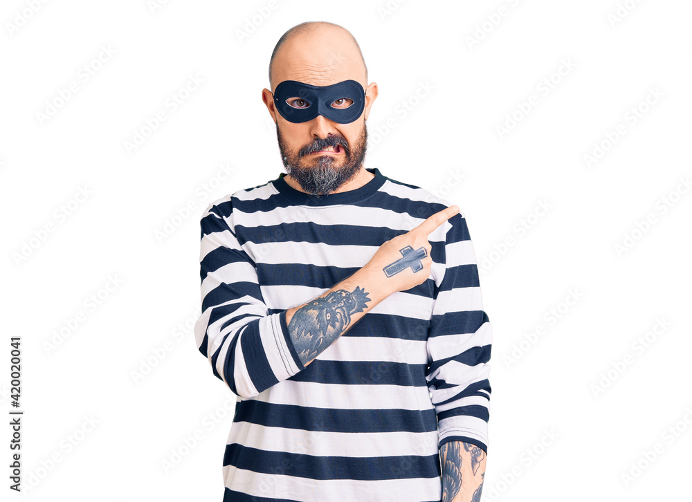 Young handsome man wearing burglar mask pointing aside worried and nervous with forefinger, concerned and surprised expression Young handsome man wearing burglar mask pointing aside worried and nervous with forefinger, concerned and surprised expression