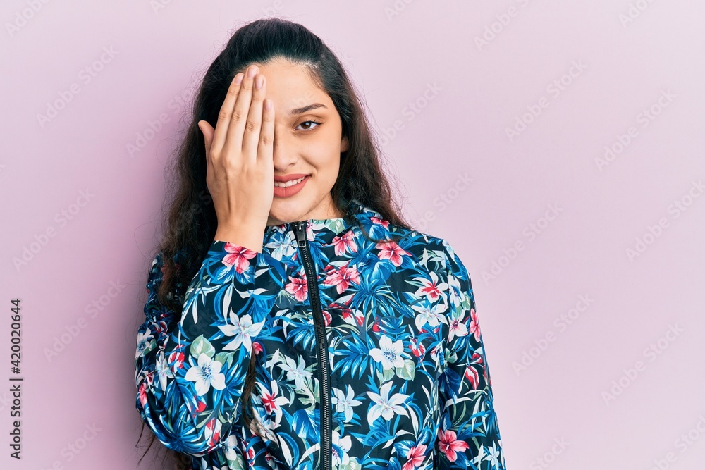 Beautiful middle eastern woman wearing casual floral jacket covering one eye with hand, confident smile on face and surprise emotion. Beautiful middle eastern woman wearing casual floral jacket covering one eye with hand, confident smile on face and surprise emotion.