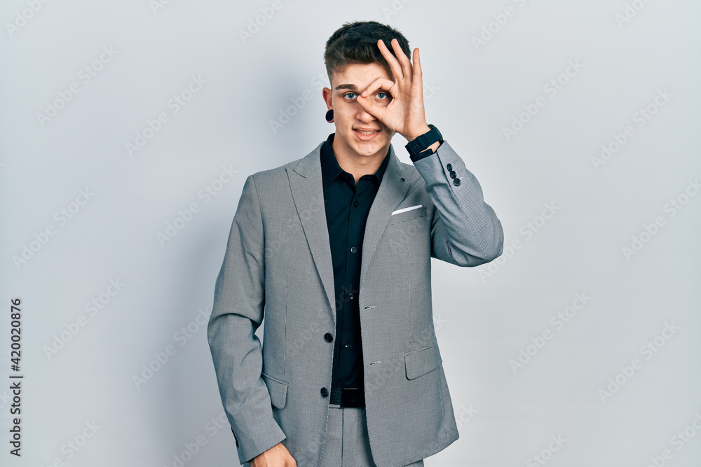 Young caucasian boy with ears dilation wearing business jacket doing ok gesture with hand smiling, eye looking through fingers with happy face. Young caucasian boy with ears dilation wearing business jacket doing ok gesture with hand smiling, eye looking through fingers with happy face.
